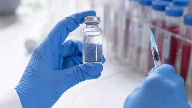 A gloved researcher holds a small glass vial of clear liquid beside a syringe on a laboratory bench, with racks of sample tubes blurred in the background.
