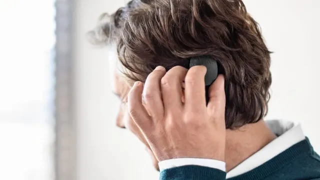 A person seen from behind and to the side reaching up to touch a small round black audio processor held against the side of the head under dark hair.