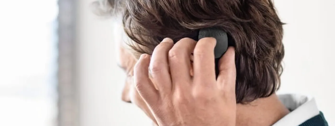 A person seen from behind and to the side reaching up to touch a small round black audio processor held against the side of the head under dark hair.