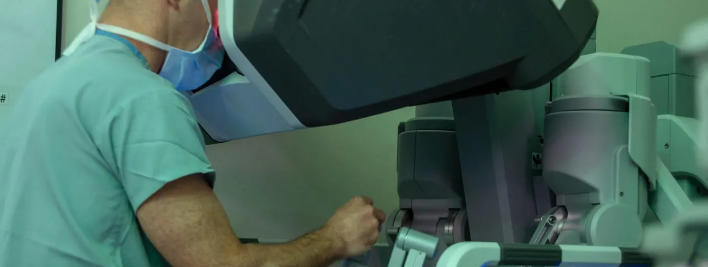 A masked surgeon in scrubs leans into the viewfinder of a robotic surgery console, hands on the controls, with the robot's articulated arms standing beside him in a teal-lit operating room.