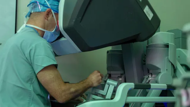 A masked surgeon in scrubs leans into the viewfinder of a robotic surgery console, hands on the controls, with the robot's articulated arms standing beside him in a teal-lit operating room.