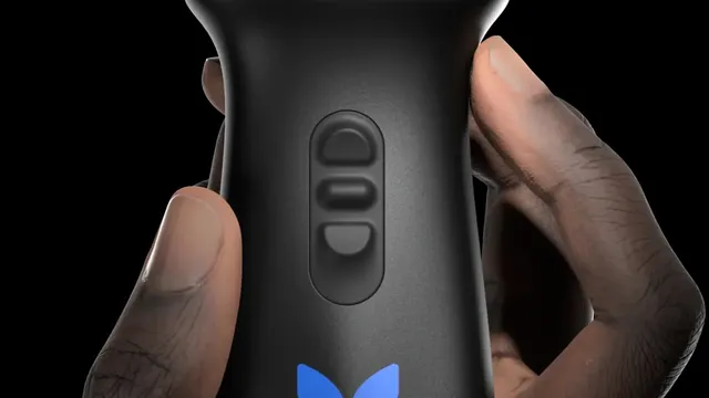 A hand holds a black handheld ultrasound probe against a dark background, its control buttons and the blue butterfly mark on the casing facing the camera.