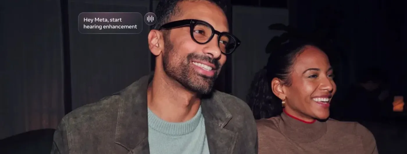 A man wearing black-framed Meta AI glasses smiles at an evening gathering as an on-screen caption reads "Hey Meta, start hearing enhancement" next to a voice waveform icon.