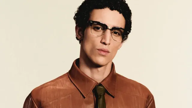 A man wearing black-and-gold Ray-Ban Meta Audio Clubmaster-style camera-free smart glasses, photographed against a plain cream backdrop.