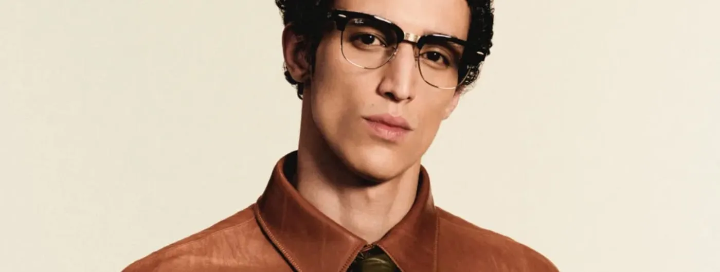 A man wearing black-and-gold Ray-Ban Meta Audio Clubmaster-style camera-free smart glasses, photographed against a plain cream backdrop.