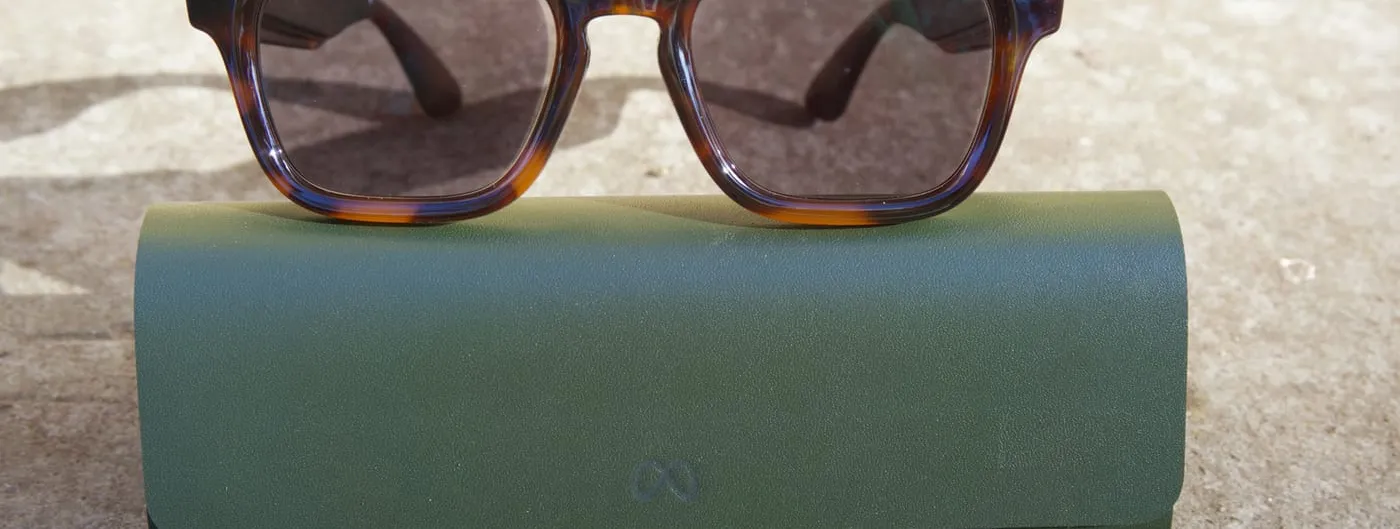 Tortoiseshell smart glasses rest on a green leather case on stone, a camera lens and a small white indicator ring visible at each corner of the frame.