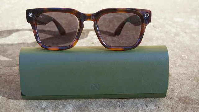 Tortoiseshell smart glasses rest on a green leather case on stone, a camera lens and a small white indicator ring visible at each corner of the frame.