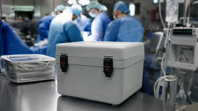 A sealed white organ transport cooler sits on a stainless steel table in an operating theatre, with gowned and masked surgical staff working out of focus behind it.