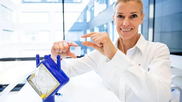 A researcher in a white shirt smiles at the camera in a bright glass-walled building, holding a small translucent rectangular membrane sample between the fingers of both hands, with a blue and yellow oxygenator cartridge standing on the bench in front of her.
