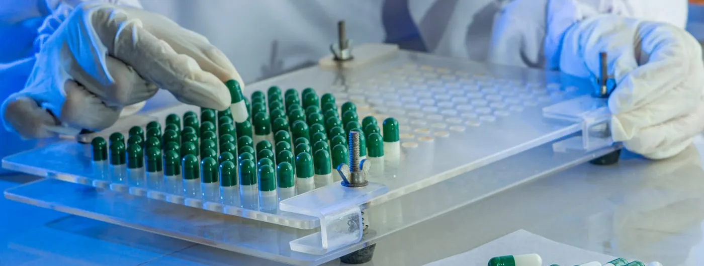 Two gloved hands work a clear acrylic capsule-filling tray holding rows of dark green and white gelatin capsules, with a small pile of finished capsules resting on paper beside it.