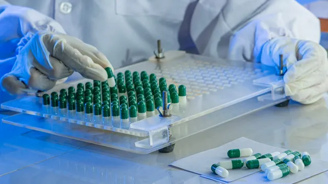 Two gloved hands work a clear acrylic capsule-filling tray holding rows of dark green and white gelatin capsules, with a small pile of finished capsules resting on paper beside it.
