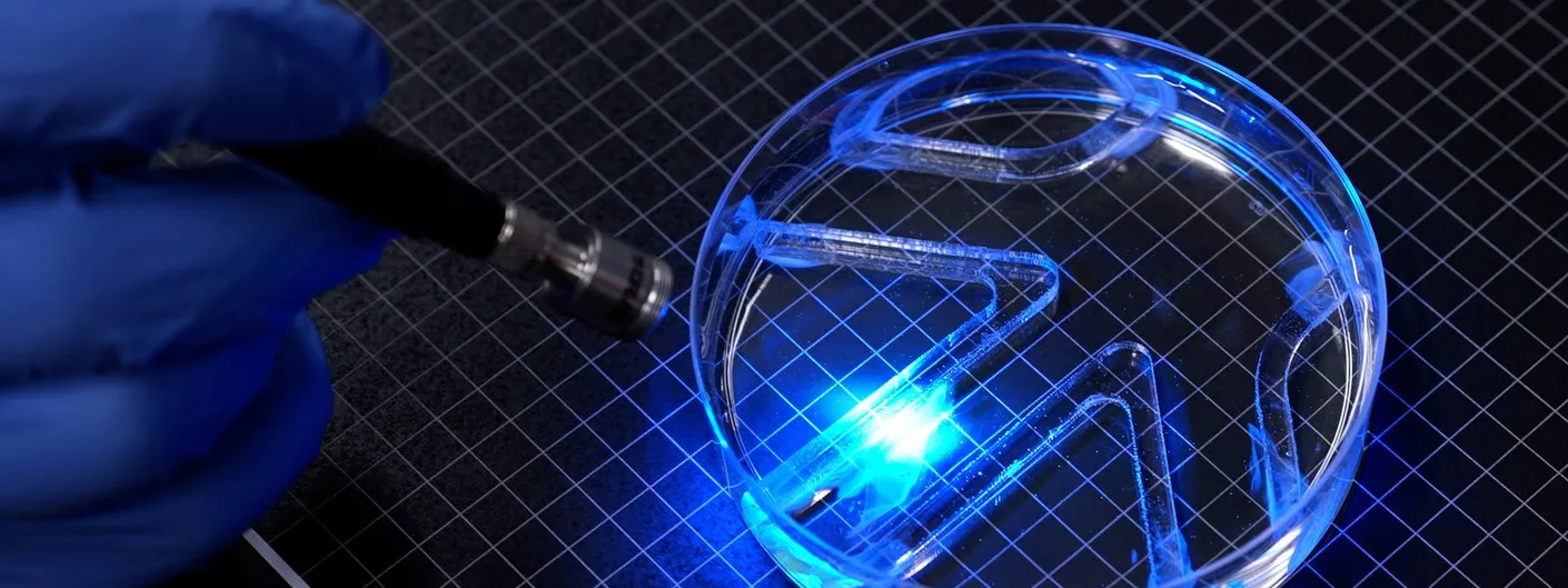 A gloved hand points a thin blue light probe at a petri dish on a grid mat; inside the dish, clear barriers form a small maze and blue light glows where the light hits the water.