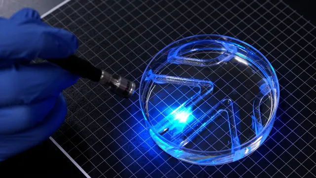 A gloved hand points a thin blue light probe at a petri dish on a grid mat; inside the dish, clear barriers form a small maze and blue light glows where the light hits the water.