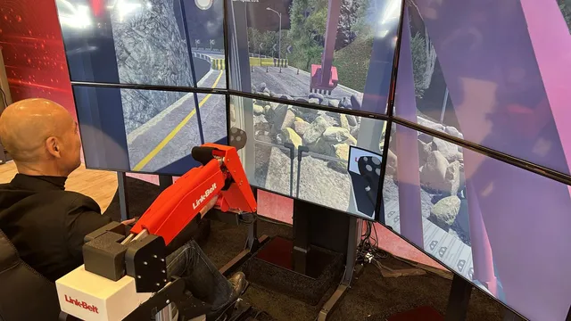A man in dark clothing sits gripping a red miniature excavator boom and bucket mounted on a chair rig, facing a wraparound array of six screens showing a virtual excavator digging among boulders on a mountain road.