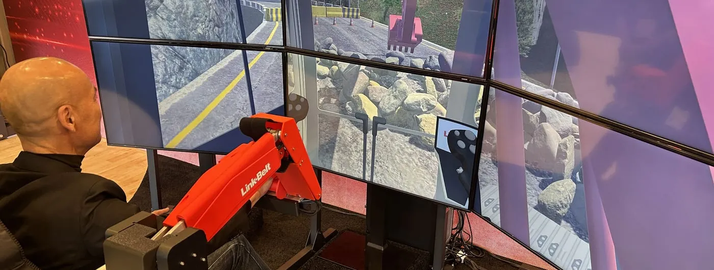A man in dark clothing sits gripping a red miniature excavator boom and bucket mounted on a chair rig, facing a wraparound array of six screens showing a virtual excavator digging among boulders on a mountain road.