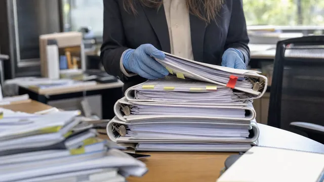 A gloved hand reviewing a tall stack of printed clinical-trial binders and tabbed folders on an office desk, one folder marked with a red flag tab.