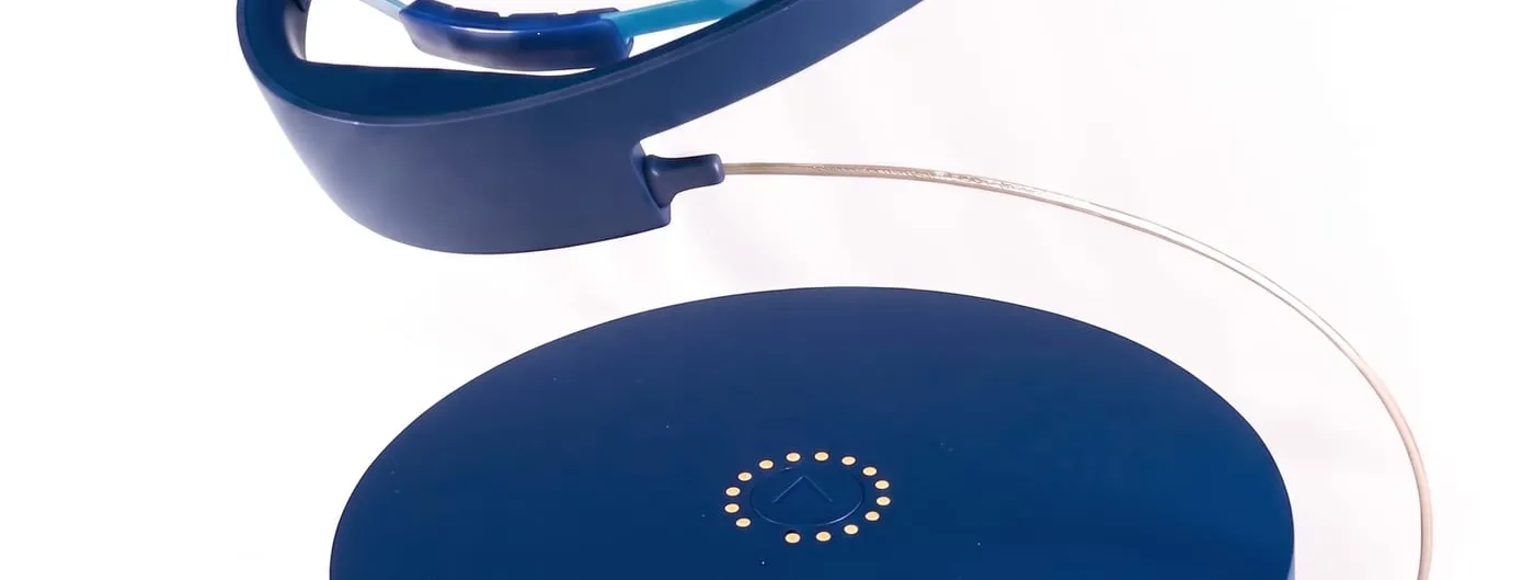 A navy-blue, lightweight head-worn magnetic therapy ring with light-blue internal contact pads, connected by a thin cable to a round tabletop console, photographed on a white studio background.
