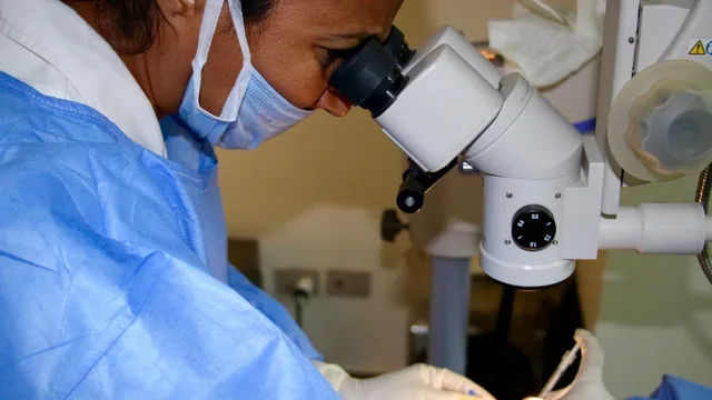 A surgeon in a face mask looks through the binocular eyepieces of an operating microscope, gloved hands holding a fine instrument at the eye of a draped patient during an intravitreal procedure.