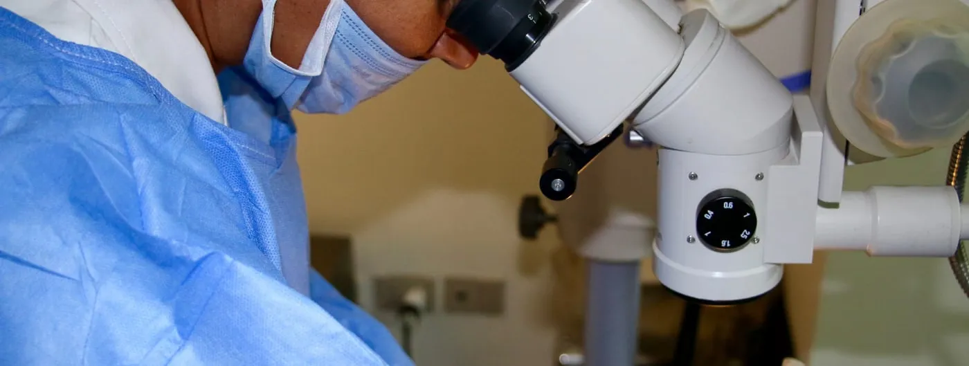 A surgeon in a face mask looks through the binocular eyepieces of an operating microscope, gloved hands holding a fine instrument at the eye of a draped patient during an intravitreal procedure.