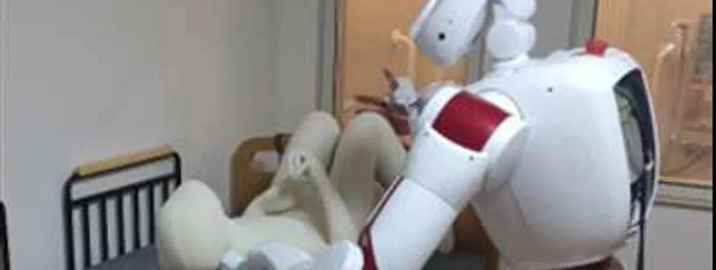 A white and red humanoid robot leans over a hospital bed, both arms under a pale mannequin, lifting it from lying flat towards a sitting position.