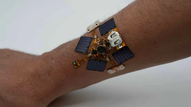 A small circuit assembly with five dark photovoltaic panels, a coin cell and a round haptic motor sits directly on the skin of a forearm, connected by orange serpentine traces.