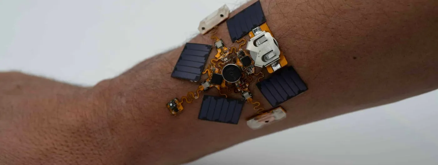 A small circuit assembly with five dark photovoltaic panels, a coin cell and a round haptic motor sits directly on the skin of a forearm, connected by orange serpentine traces.