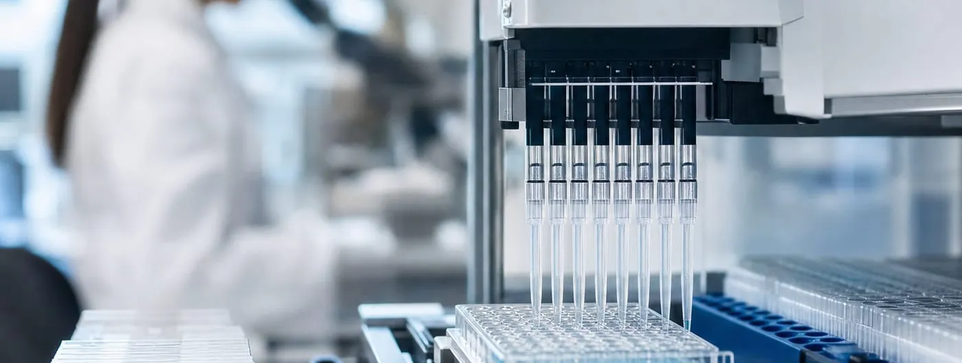 An automated pipetting head lowers eight tips into a clear multi-well plate on a laboratory bench, a researcher in a white coat out of focus behind it.