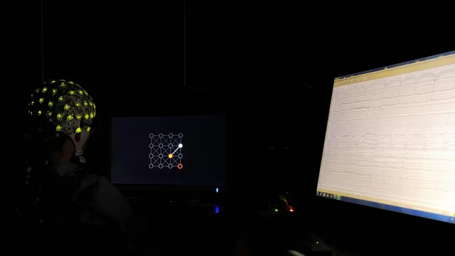 A participant wears a green-electrode EEG cap while a nearby monitor displays a live scrolling EEG waveform trace during a brain-computer interface experiment.