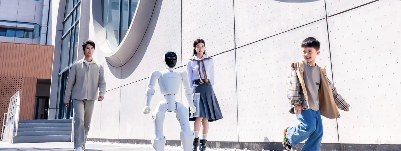 A white NOETIX-branded bipedal humanoid robot with a black visor-style head walks outdoors on a paved plaza past a man, a girl in a school uniform and a running boy, in front of a curved concrete building facade.