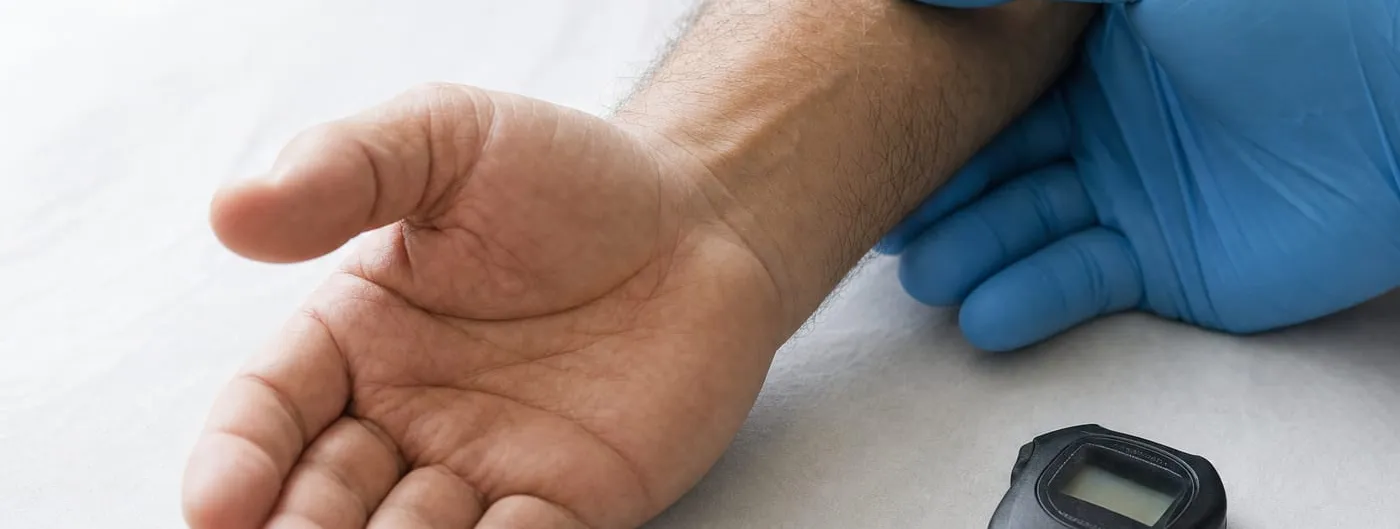 A close view of an adult hand resting palm-up on a clinic examination table while a gloved hand supports the wrist, with a plain stopwatch lying beside it.