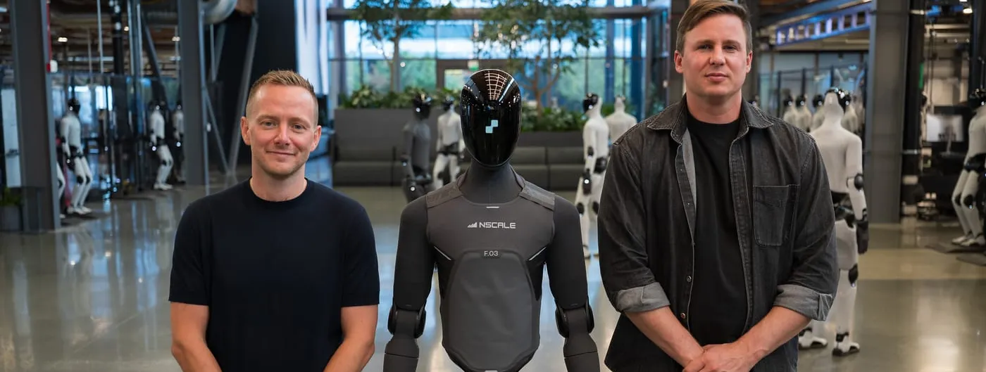 Two men in dark clothing stand on either side of a black humanoid robot wearing an Nscale-branded torso panel, inside a bright robotics facility with rows of other humanoids behind them.