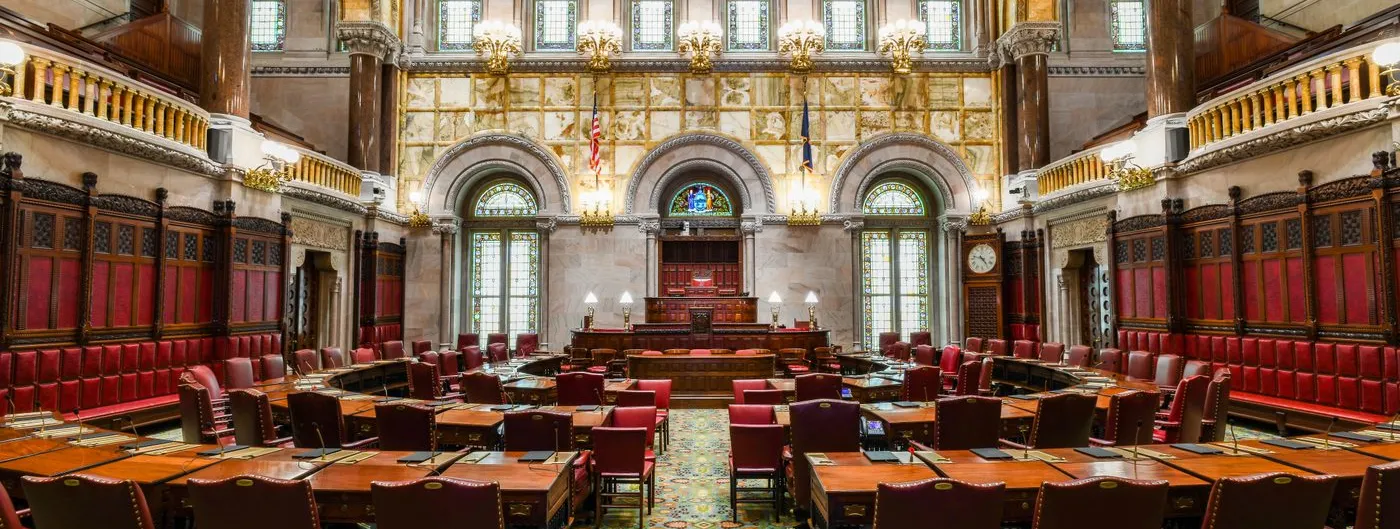 The empty New York State Senate chamber in Albany, with rows of red leather chairs at wooden desks beneath stained-glass windows and carved stone arches.
