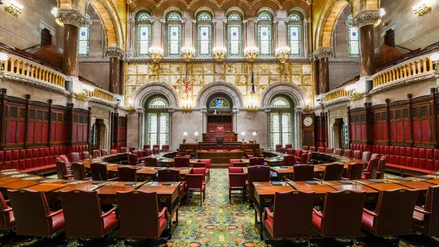 The empty New York State Senate chamber in Albany, with rows of red leather chairs at wooden desks beneath stained-glass windows and carved stone arches.