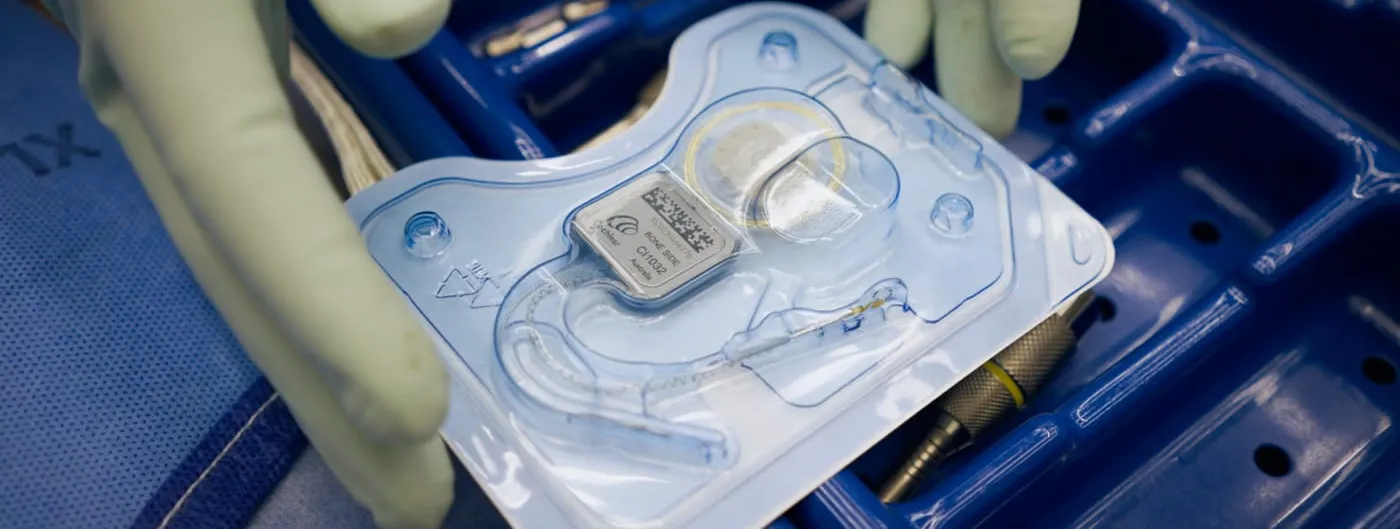 Two gloved hands lift a sealed sterile blister pack holding a cochlear implant — a small square receiver-stimulator with a coiled electrode lead — above a blue perforated surgical instrument tray.