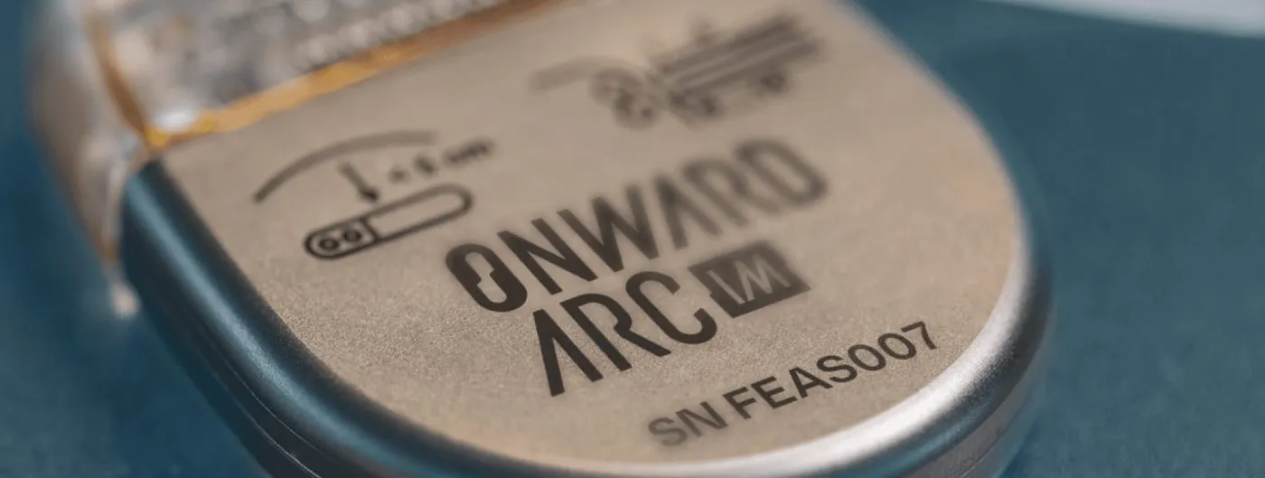Close-up of a rounded metal implantable pulse generator lying on a blue-grey surface, its titanium face laser-engraved with the ONWARD ARC IM name, a small diagram and the serial number SN FEAS007, with translucent lead connectors entering the header.