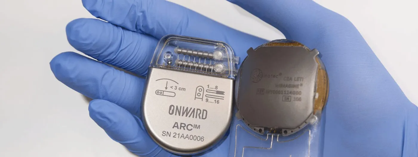 A gloved hand holds ONWARD Medical's ARC-IM implantable neurostimulator next to the paired WIMAGINE brain-computer-interface implant, showing both pieces of hardware that make up the ARC-BCI system.