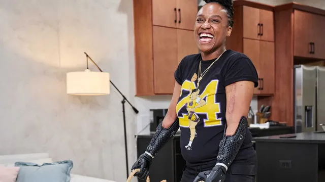 A laughing woman with a Hero PRO bionic arm on each side lifts a wicker laundry basket by its handles in a kitchen.