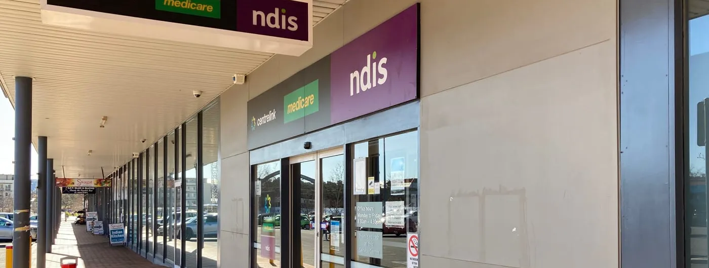 Storefront entrance of a Services Australia service centre in Canberra, with wall-mounted and hanging signs reading Centrelink, Medicare and NDIS above the glass doors.