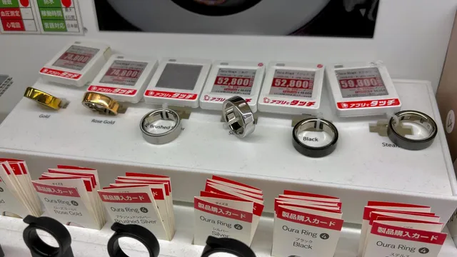 Oura smart rings in gold, rose gold and silver finishes displayed on a white retail plinth beside Japanese-language price tags and stacks of purchase cards in a Tokyo electronics shop.