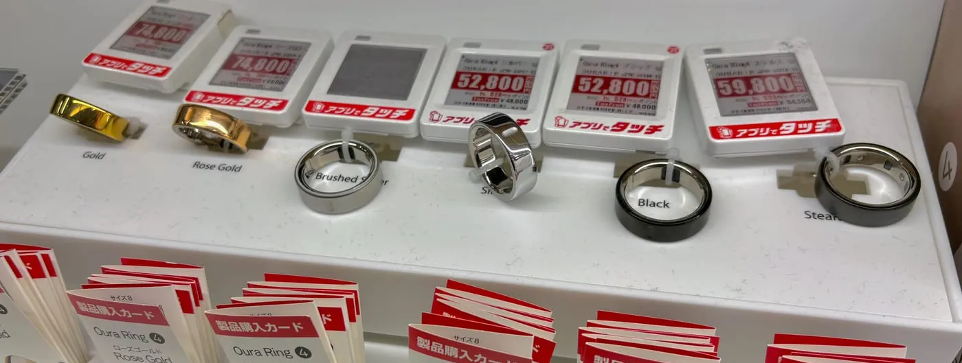 Oura smart rings in gold, rose gold and silver finishes displayed on a white retail plinth beside Japanese-language price tags and stacks of purchase cards in a Tokyo electronics shop.
