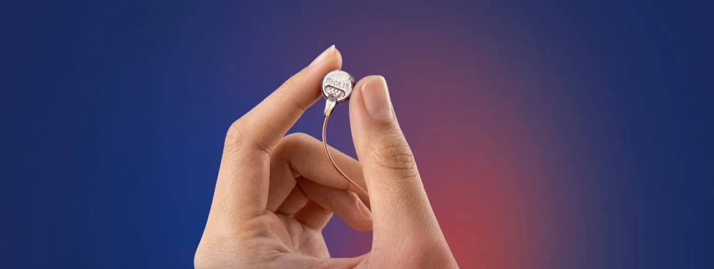 A hand holds a small round implant module between thumb and forefinger, a fine wire trailing from it, against a blue and red gradient background.