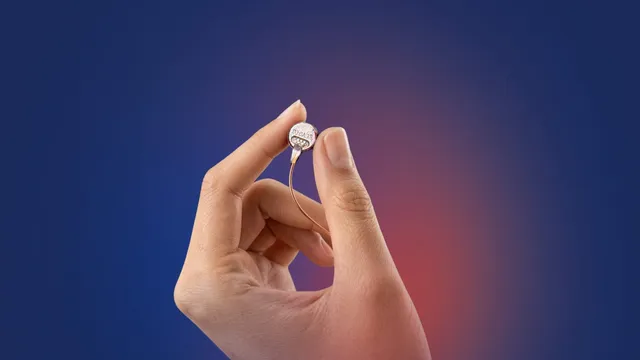 A hand holds a small round implant module between thumb and forefinger, a fine wire trailing from it, against a blue and red gradient background.