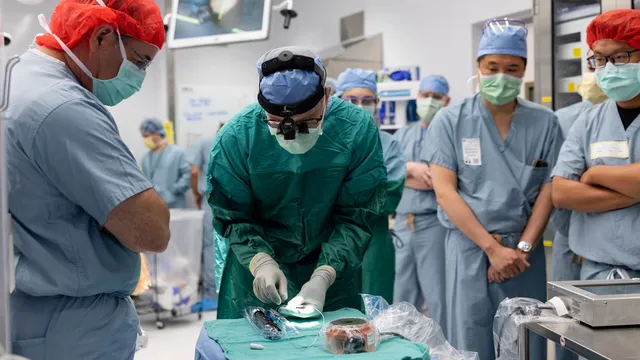 A surgeon in green scrubs and a head-mounted loupe leans over a draped instrument table handling implant components, watched by six colleagues in blue scrubs and masks in an operating room.