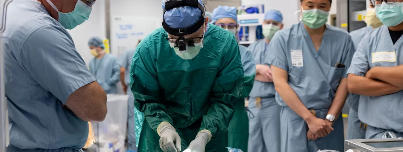 A surgeon in green scrubs and a head-mounted loupe leans over a draped instrument table handling implant components, watched by six colleagues in blue scrubs and masks in an operating room.