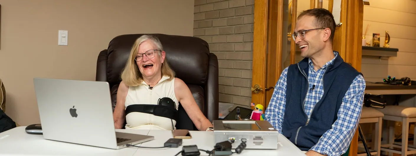 Trial participant laughs while communicating via the Connexus BCI's tabletop processing unit and a laptop, seated beside investigator Matthew Willsey in her home.