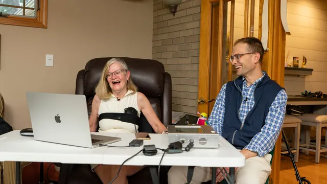 Trial participant laughs while communicating via the Connexus BCI's tabletop processing unit and a laptop, seated beside investigator Matthew Willsey in her home.