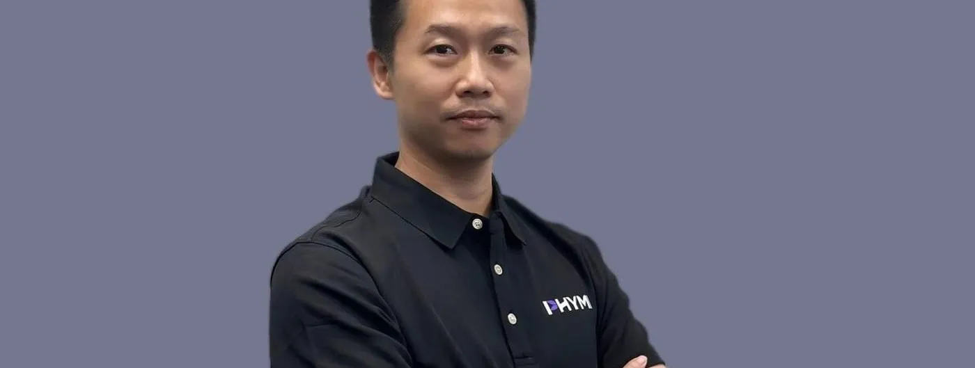 Liu Nianqiu, founder of PHYMI, stands with arms crossed wearing a black polo shirt embroidered with the PHYMI logo against a plain grey-blue background.