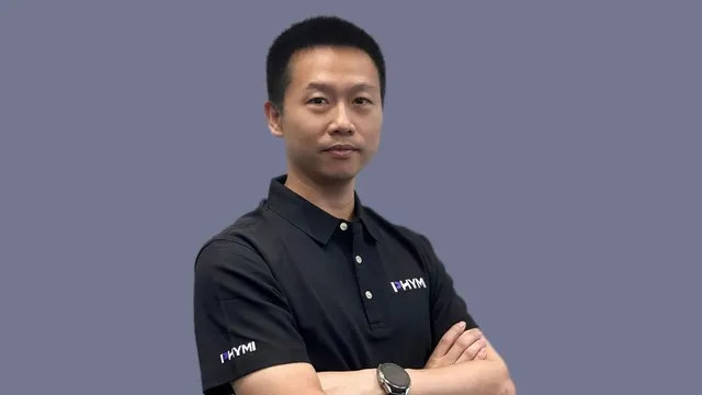 Liu Nianqiu, founder of PHYMI, stands with arms crossed wearing a black polo shirt embroidered with the PHYMI logo against a plain grey-blue background.