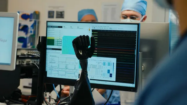 A black robotic prosthetic hand rises in front of a monitor showing live neural-signal traces and a decoding interface, with two masked, surgical-cap-wearing lab staff blurred in the background of a research lab.