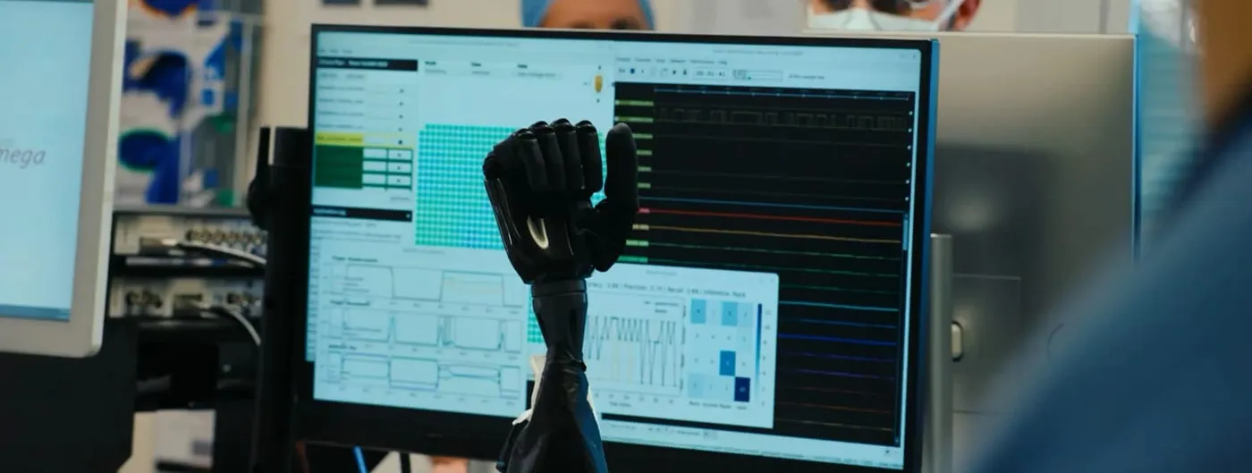 A black robotic prosthetic hand rises in front of a monitor showing live neural-signal traces and a decoding interface, with two masked, surgical-cap-wearing lab staff blurred in the background of a research lab.